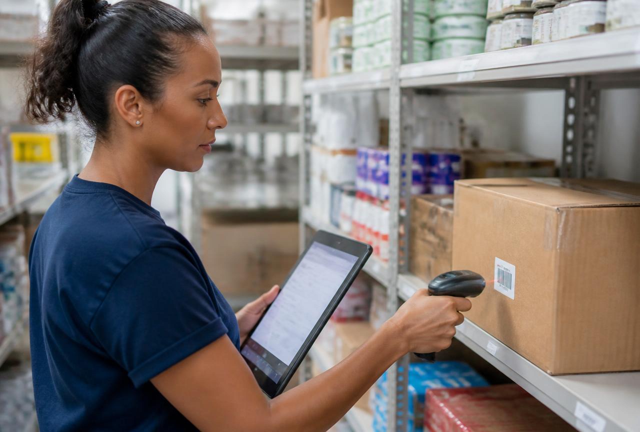 Colaboradora de pequena empresa usando tablet e leitor de código de barras para conferir caixas em prateleiras organizadas do estoque